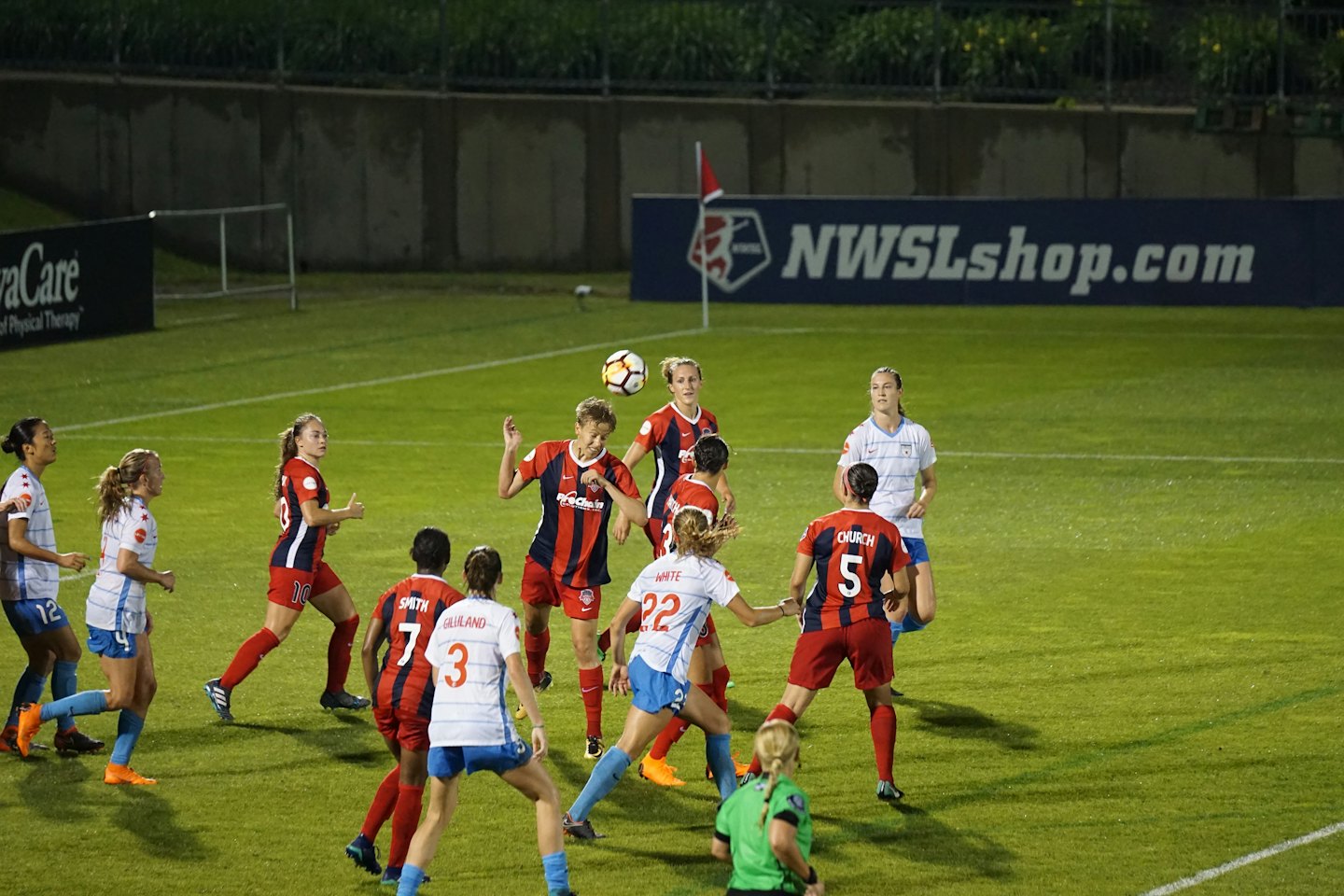 Female soccer players in red and white jerseys compete during an evening game.