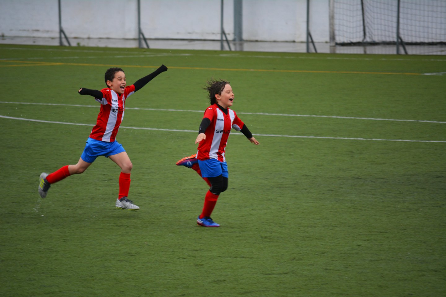 Two boys in soccer uniforms run across a field celebrating.